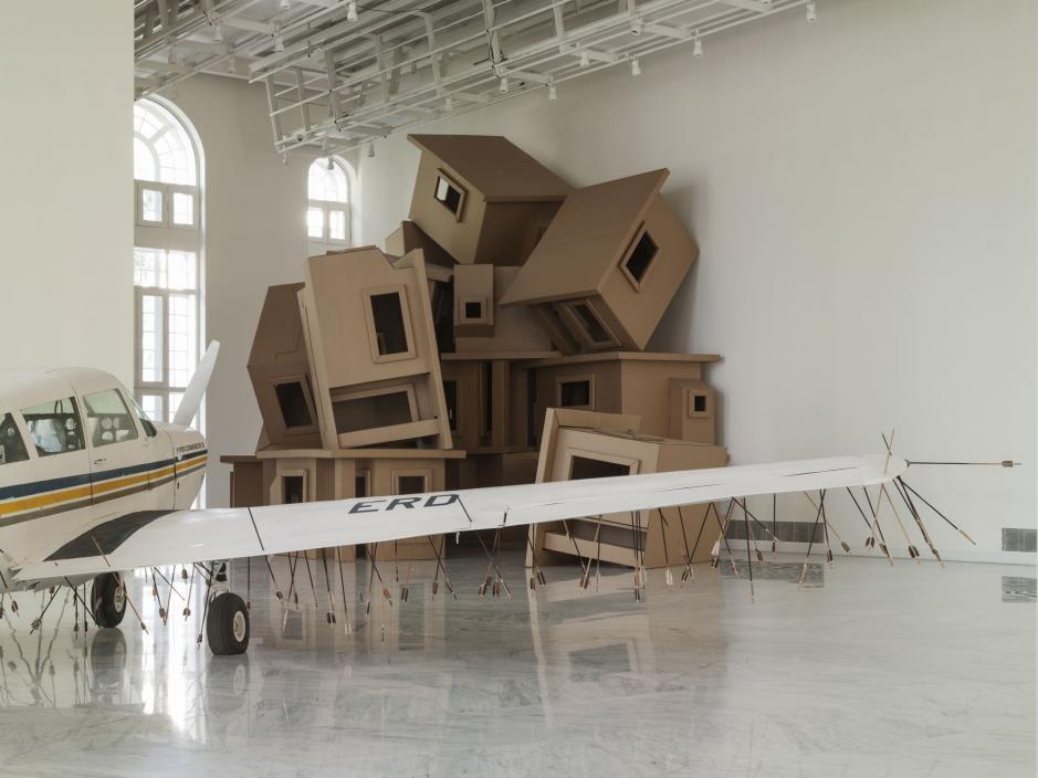 Cardboard houses stacked with a plane whose underbelly is covered in arrows in the foreground.