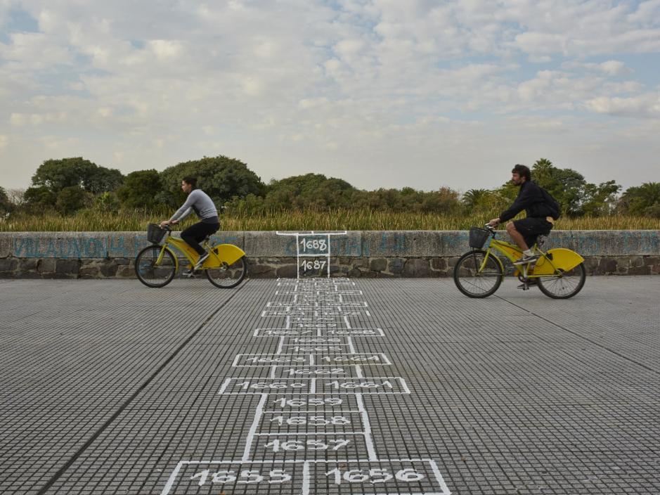 People biking over on a sidewalk that has a long hopscotch drawn on 