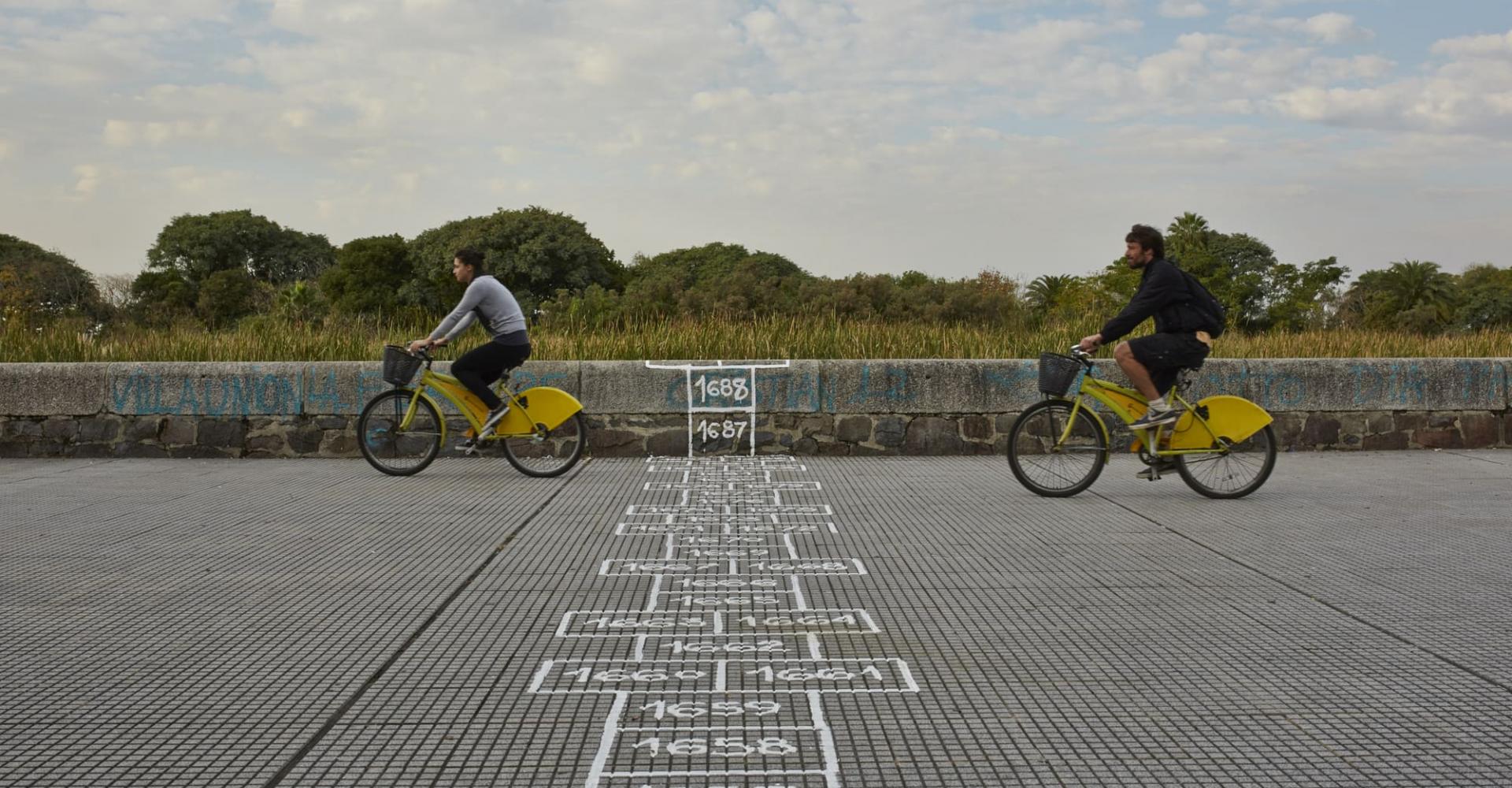 People biking over on a sidewalk that has a long hopscotch drawn on 